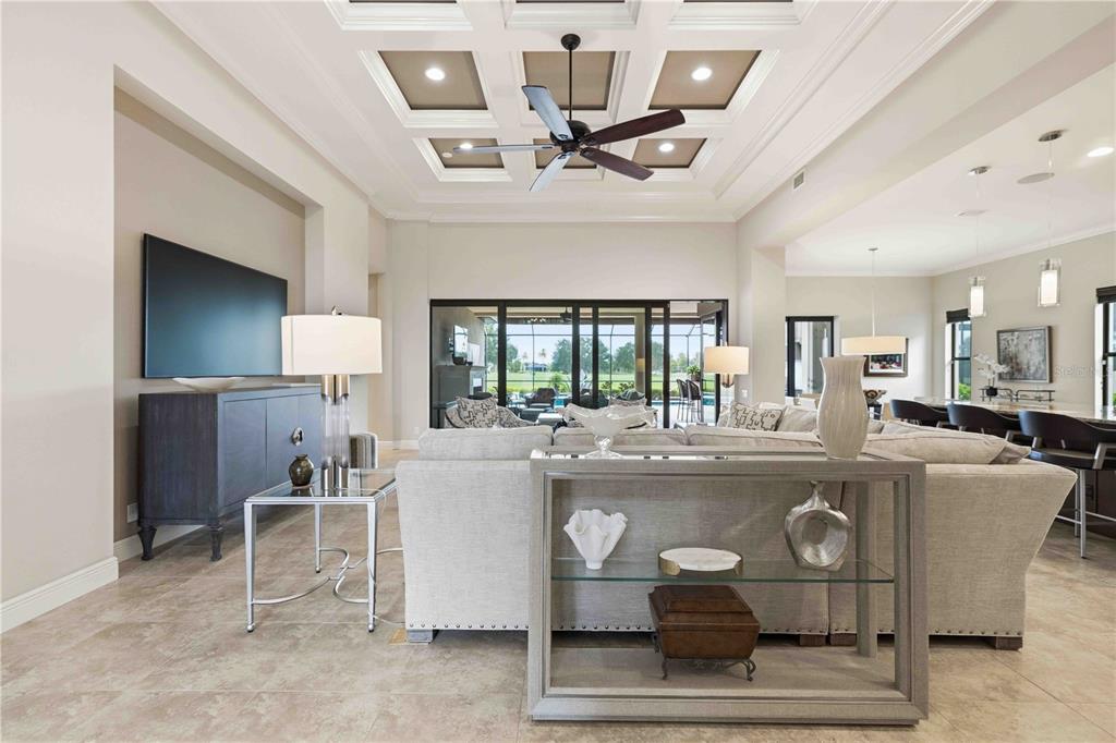 A spacious living room features a coffered ceiling, neutral furniture, and large sliding glass doors — 7638 WINDY HILL COVE, BRADENTON, FL