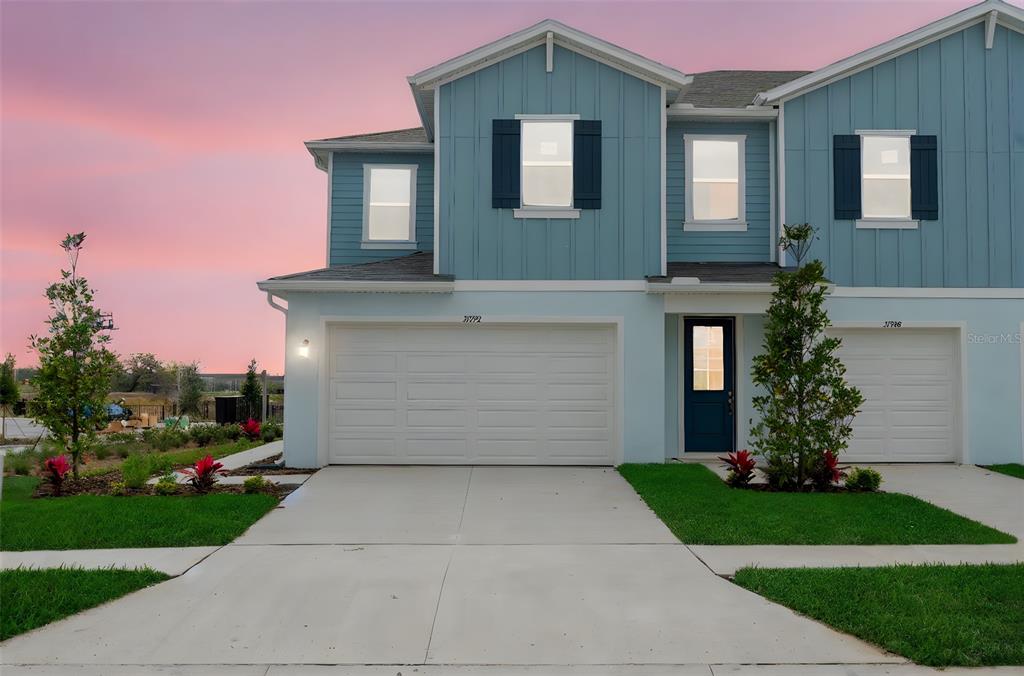 A two-story duplex with blue siding, white garage doors, and a concrete driveway at sunset — 31781 KESTREL CALLING DRIVE, SAN ANTONIO, FL