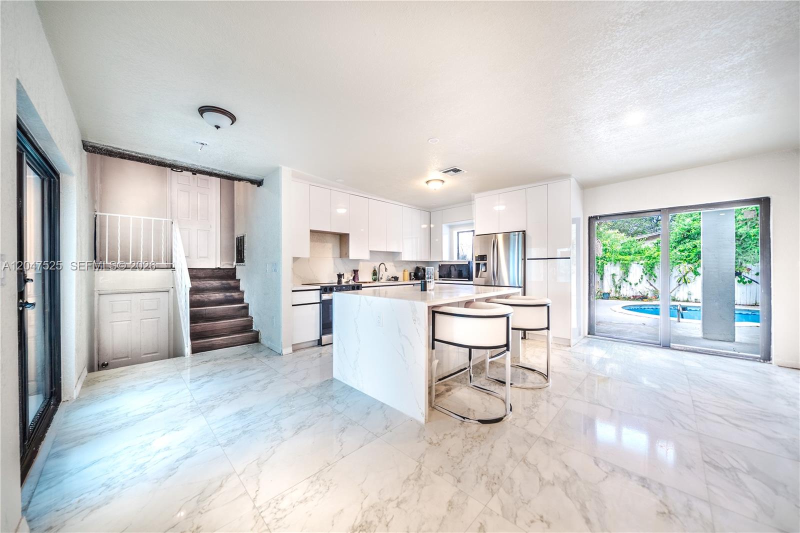 A modern kitchen features a marble island, white cabinetry, and sliding doors overlooking a pool — 20200 NE 14th Ave, Miami FL 33179, MIAMI, FL