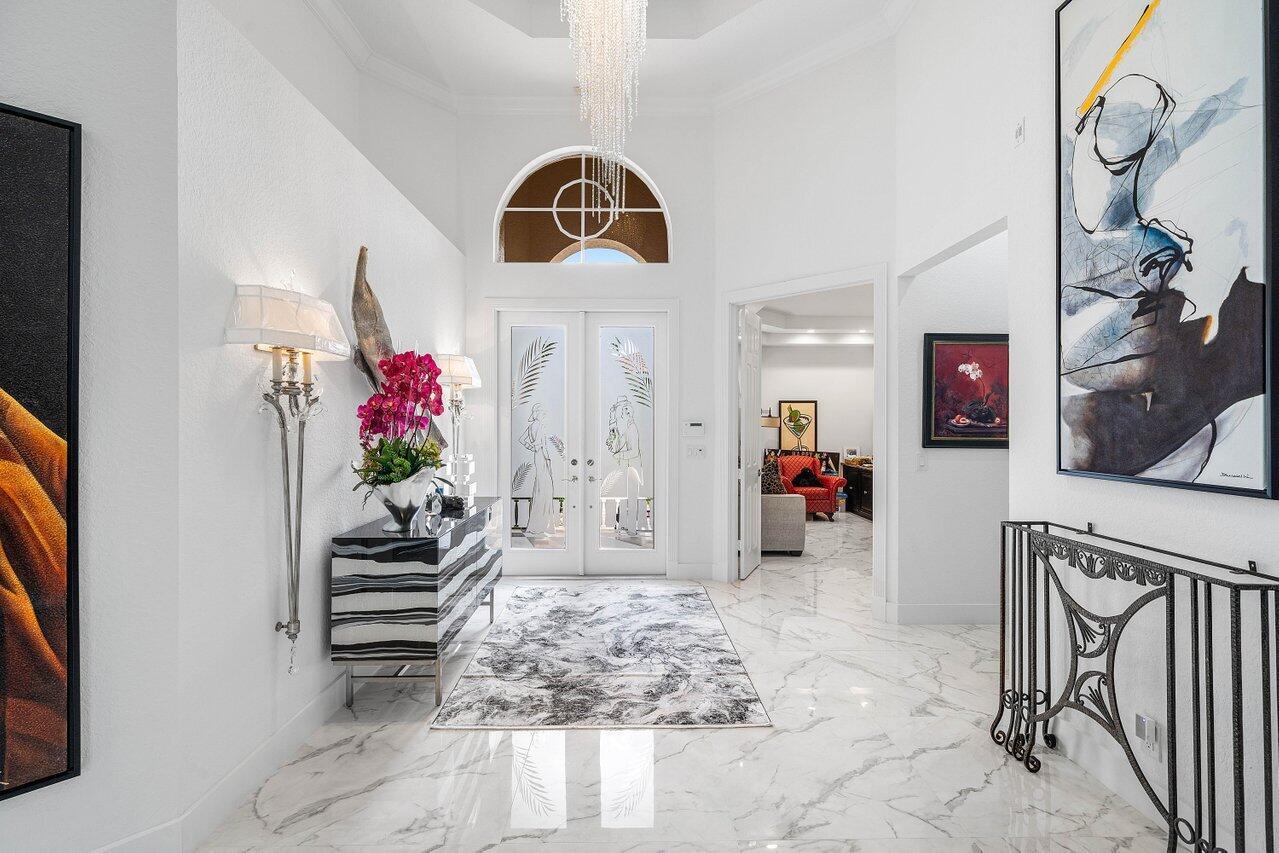 A bright, marble-floored foyer features a crystal chandelier, double glass doors, and modern wall art — 6517 Somerset Circle, Boca Raton FL 33496, BOCA RATON, FL