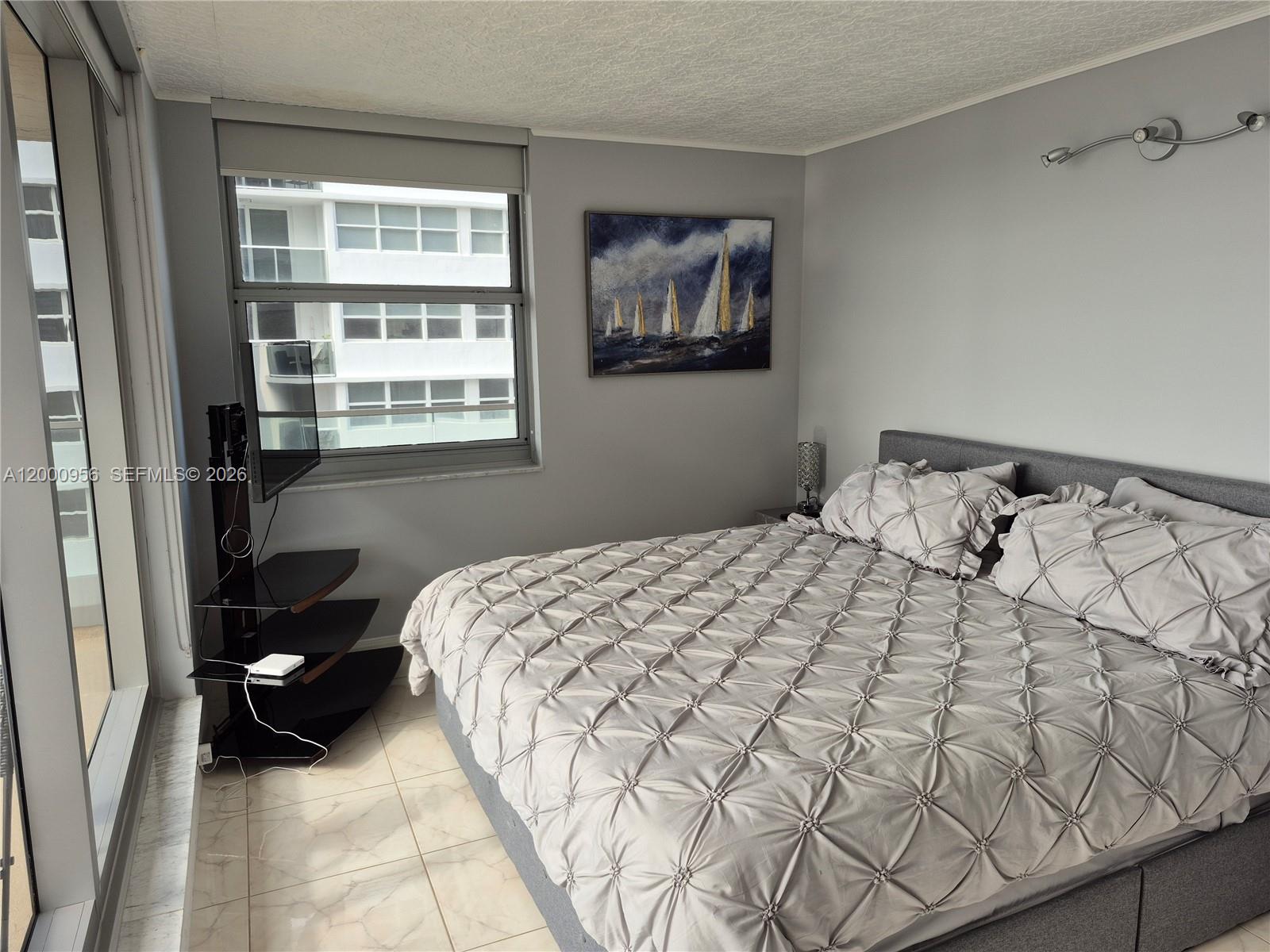 A bedroom features a gray tufted bed, a corner television stand, and a window view — 1912 S Ocean Dr # 17C, Hallandale Beach FL 33009, HALLANDALE BEACH, FL