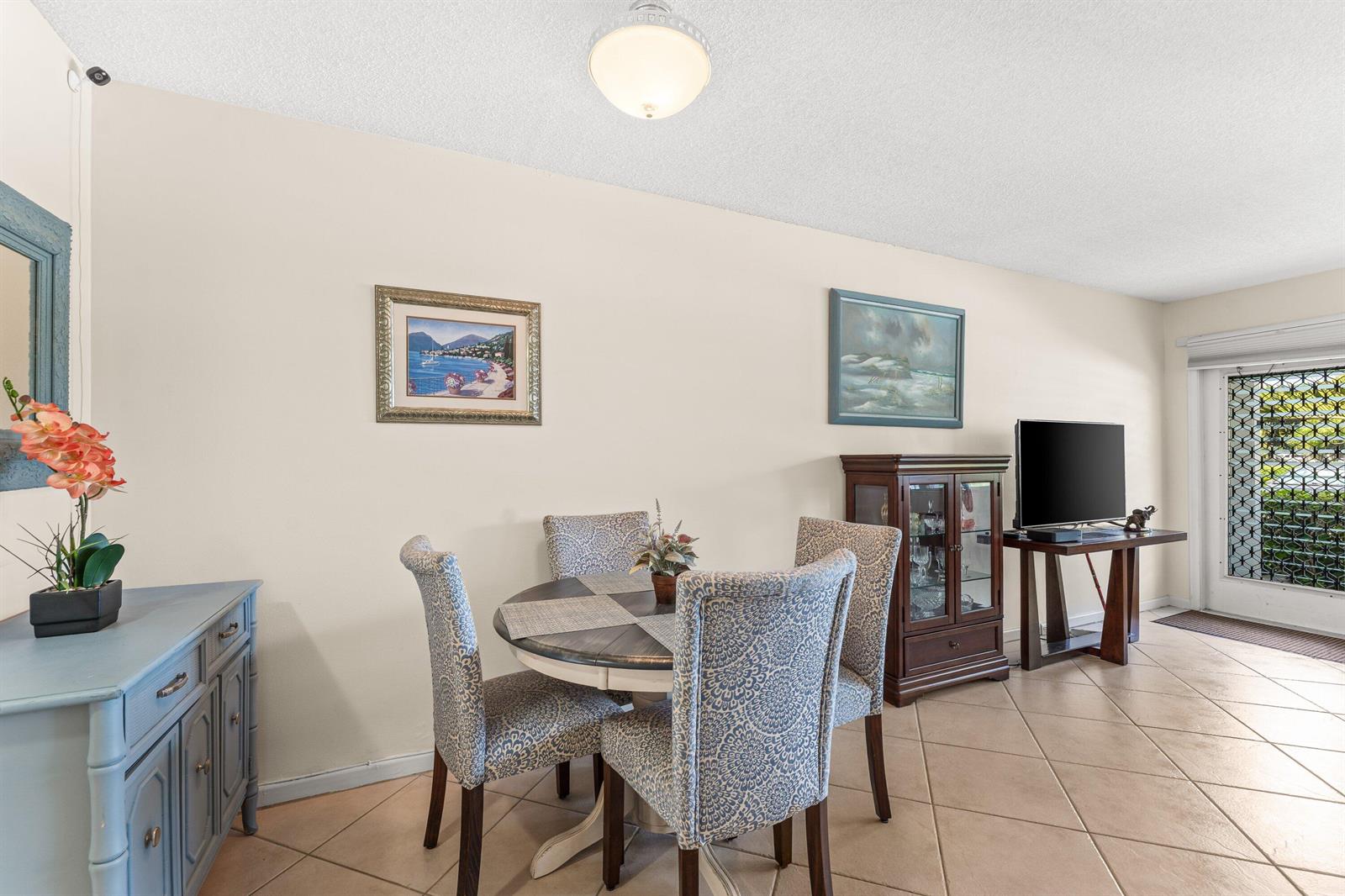 A dining area features a round table with four patterned chairs, tile flooring, and furniture — 1017 Harwood C, Deerfield Beach FL 33442, DEERFIELD BEACH, FL