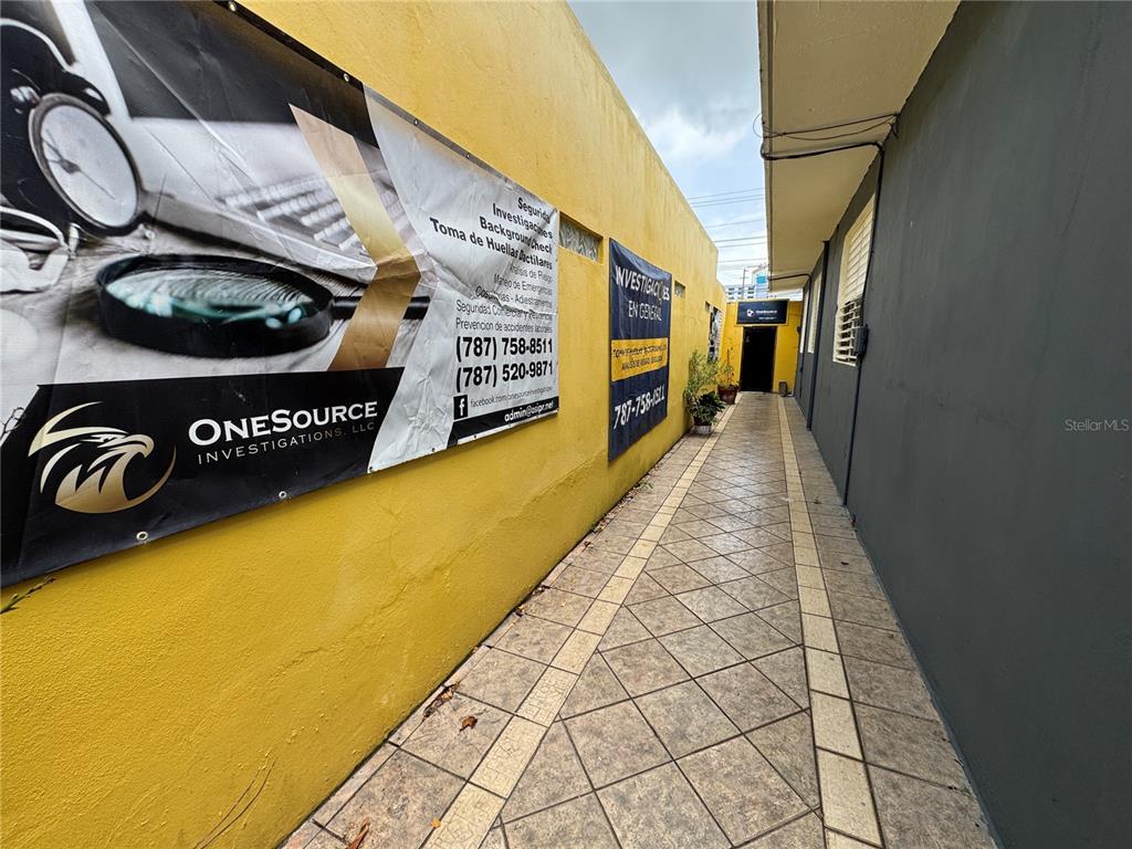 A narrow outdoor walkway leads to a building entrance with large investigative service banners displayed — #339 COLL Y TOSTE, SAN JUAN, PR