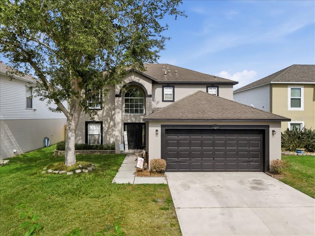 A two-story house with a beige stucco exterior, a dark garage door, and a large tree — 1038 MACON DRIVE, TITUSVILLE, FL