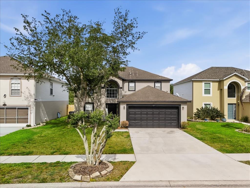 A two-story suburban house with a dark garage door and a large tree in front — 1038 MACON DRIVE, TITUSVILLE, FL