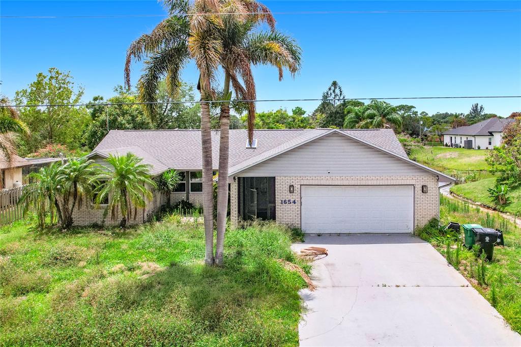 A single-story house with a brick and siding exterior, a two-car garage, and overgrown landscaping — 1654 SE FALLON DRIVE, PORT ST LUCIE, FL