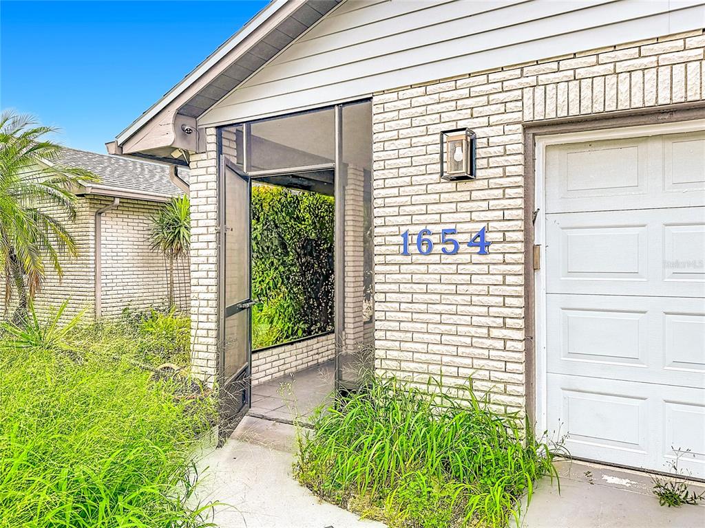 A brick house exterior features a screened entryway, a white garage door, and overgrown landscaping — 1654 SE FALLON DRIVE, PORT ST LUCIE, FL