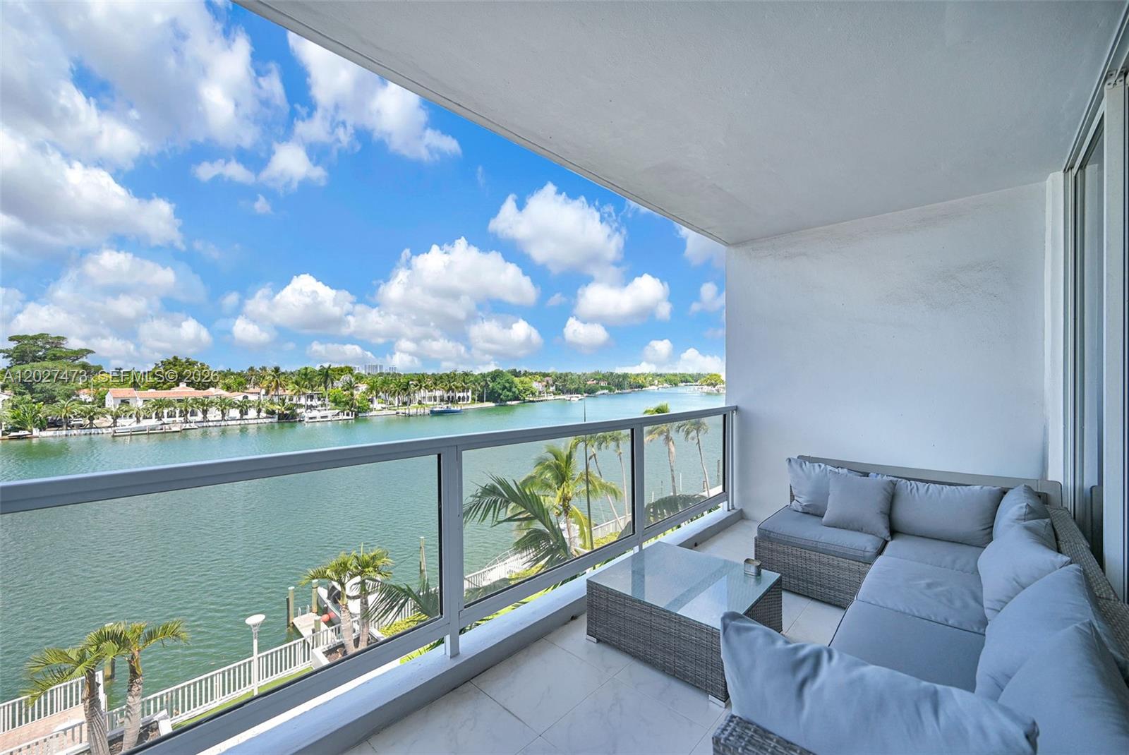 A balcony with gray wicker furniture overlooks a canal lined with palm trees and houses — 5700 Collins Ave # 5J, Miami Beach FL 33140, MIAMI BEACH, FL