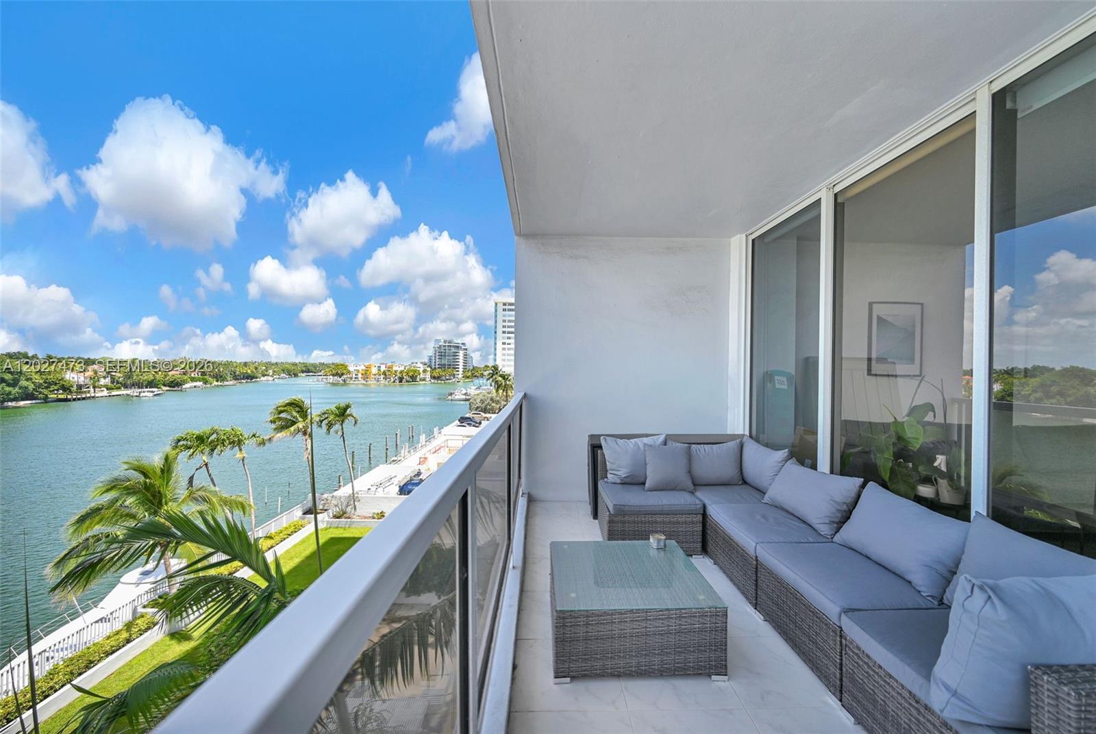 A balcony with gray sectional seating and a glass-topped table overlooks a waterfront view — 5700 Collins Ave # 5J, Miami Beach FL 33140, MIAMI BEACH, FL