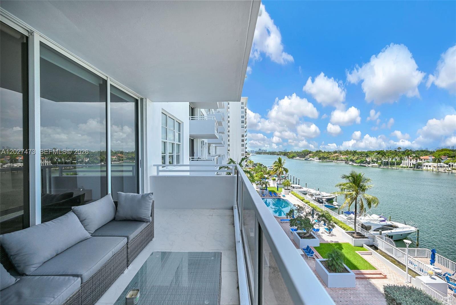 A balcony with gray outdoor furniture overlooks a swimming pool, a waterway, and boats — 5700 Collins Ave # 5J, Miami Beach FL 33140, MIAMI BEACH, FL