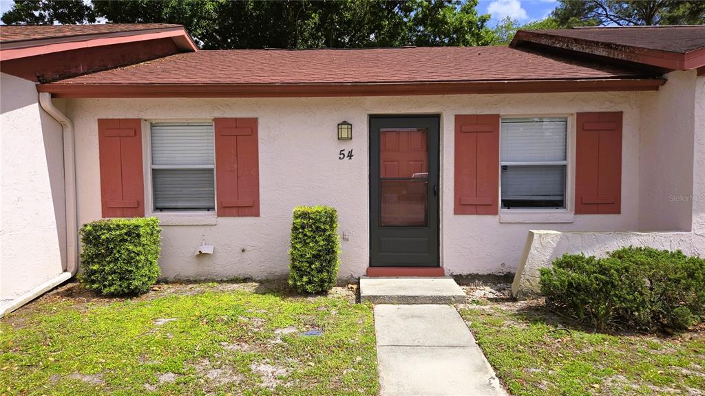 The exterior of a single-story home features a front door, two windows with shutters, and landscaping — 54 W MARBRISA WAY, KISSIMMEE, FL