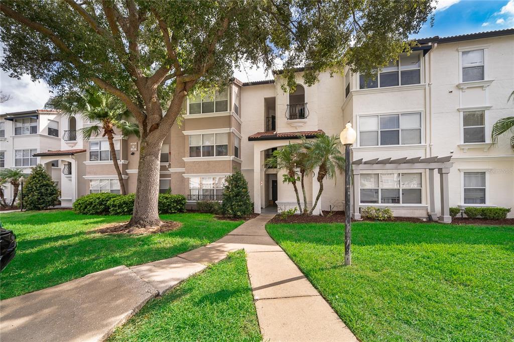 A multi-story apartment building exterior with a concrete walkway, large tree, and green lawn — 5124 CONROY ROAD UNIT 26, ORLANDO, FL