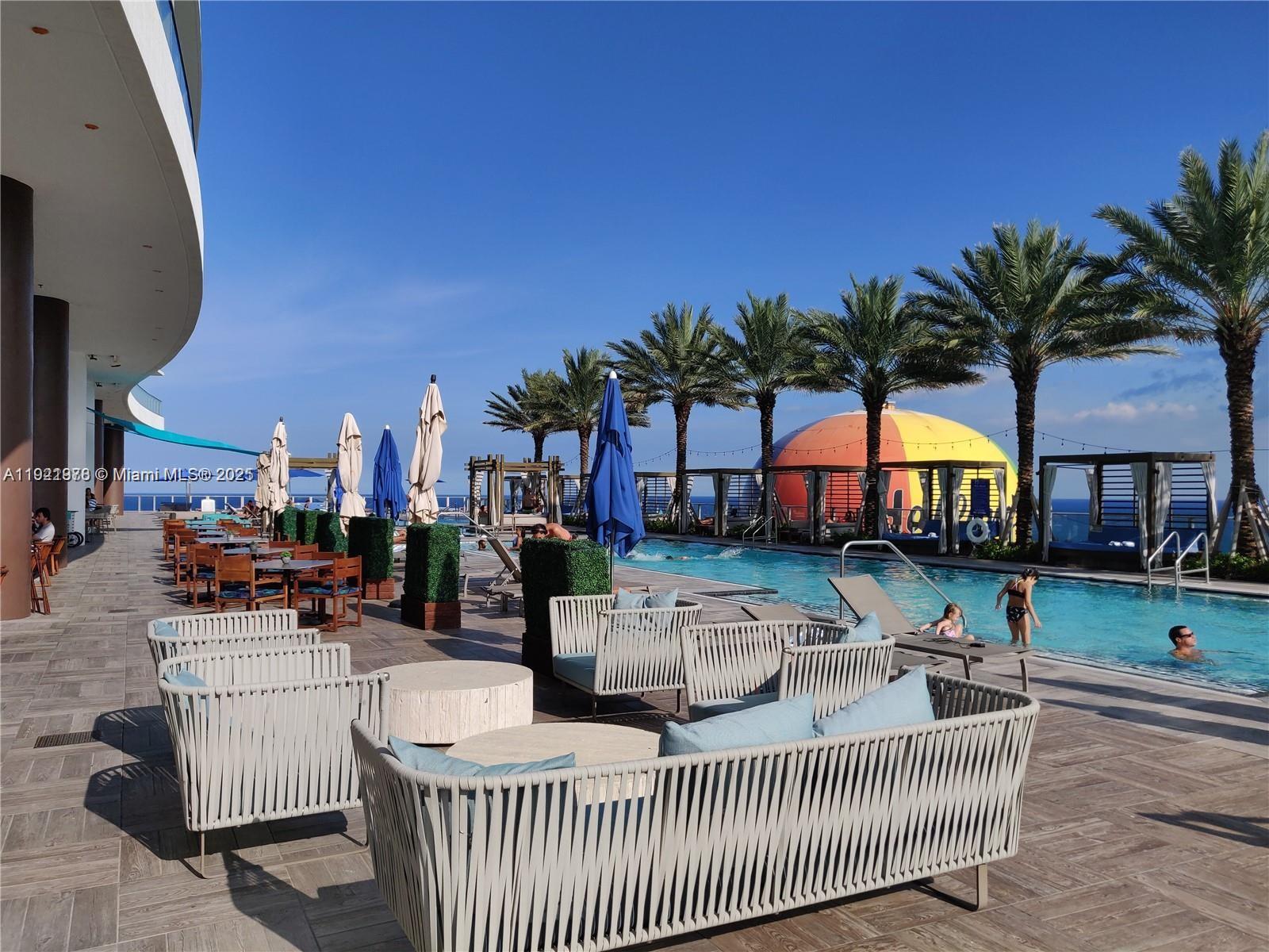 An outdoor pool deck features lounge seating, dining tables, umbrellas, and palm trees by the ocean — 4111 S Ocean Dr # 1506, Hollywood FL 33019, HOLLYWOOD, FL