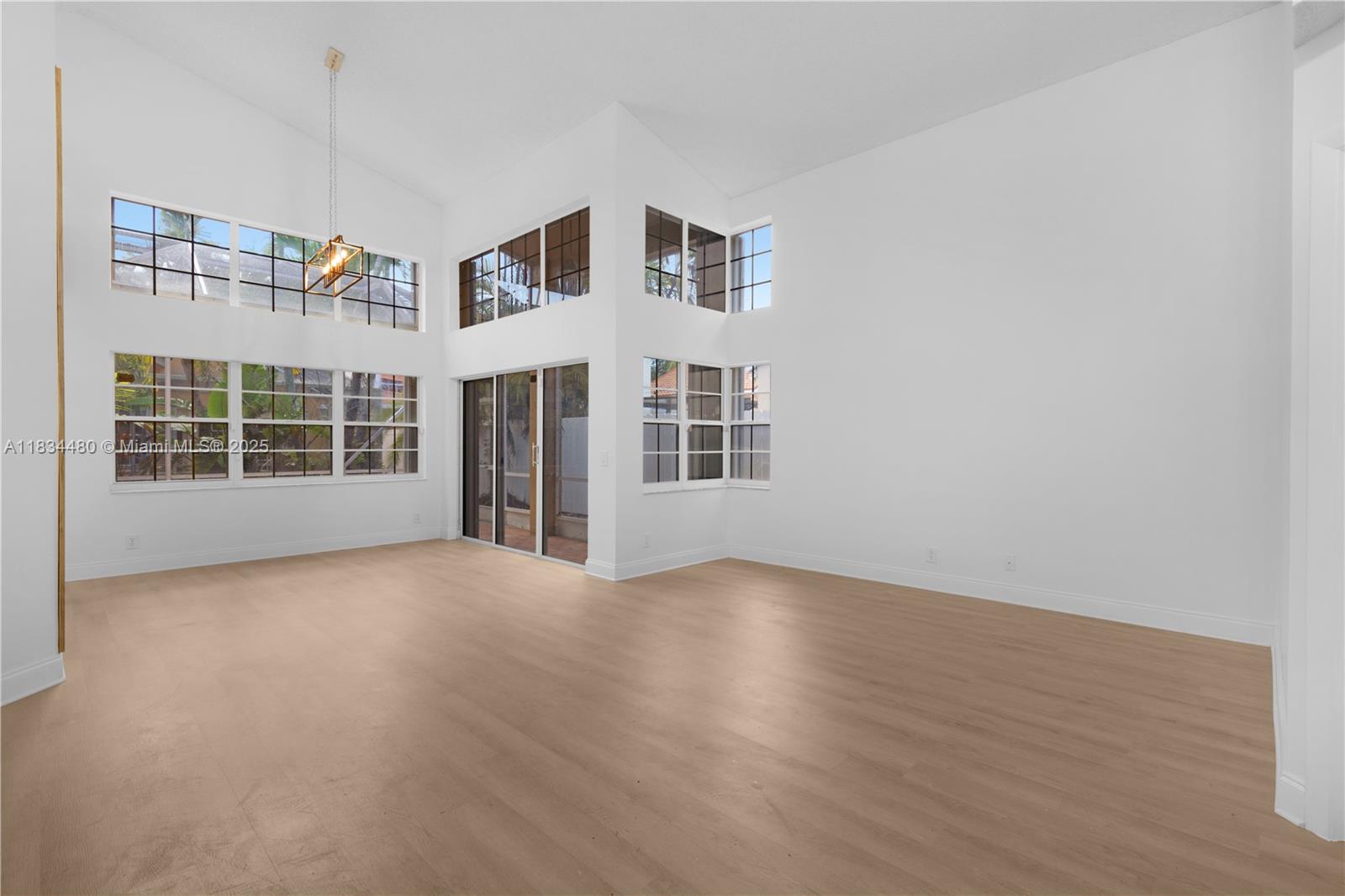 A bright, empty living room features high vaulted ceilings, large windows, and light wood flooring — 1525 Seabay Rd, Weston FL 33326, WESTON, FL