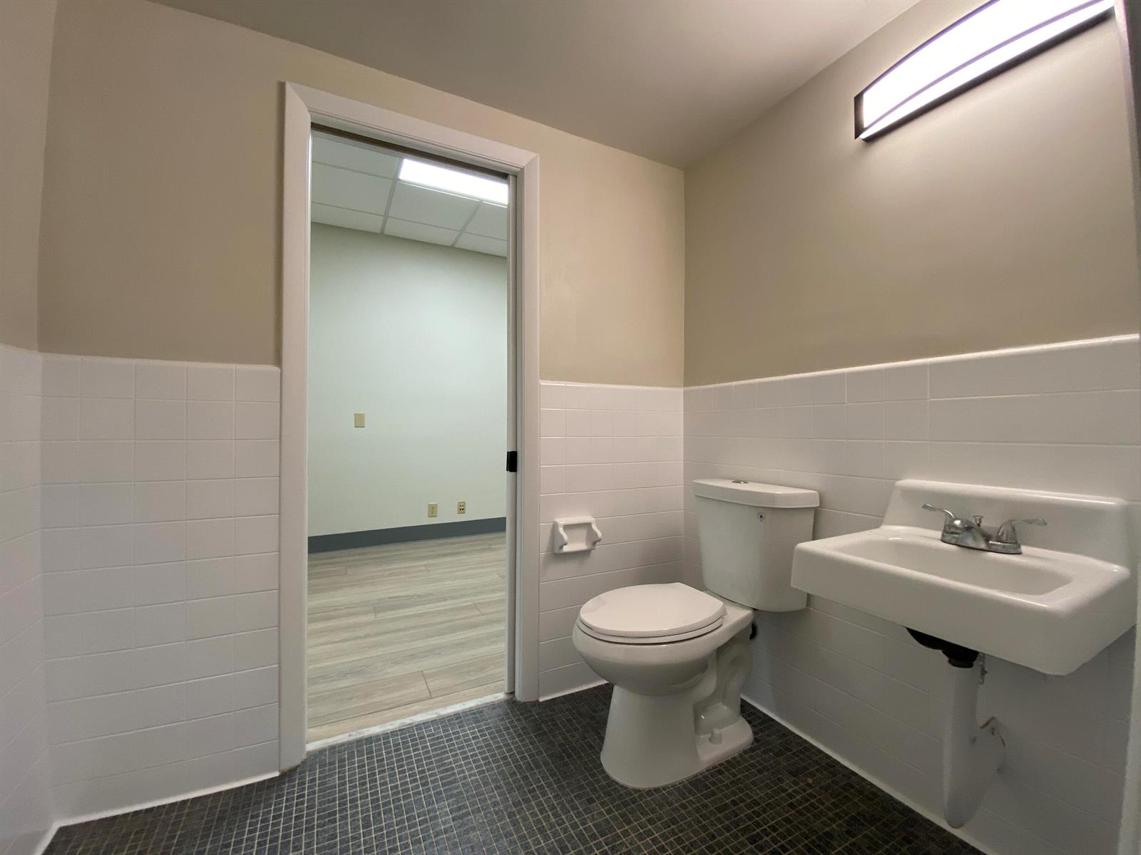 A small bathroom with a toilet, wall-mounted sink, tiled walls, and a doorway to an office — 2231 NE 25th Avenue # 1, Pompano Beach FL 33062, POMPANO BEACH, FL
