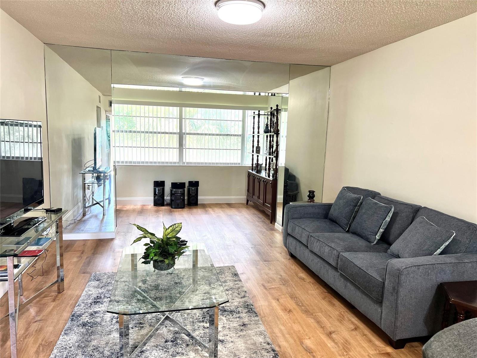 A living room features a gray sofa, glass coffee table, wood-look flooring, and mirrored walls — 3080 Holiday Springs Boulevard # 301, Margate FL 33063, MARGATE, FL