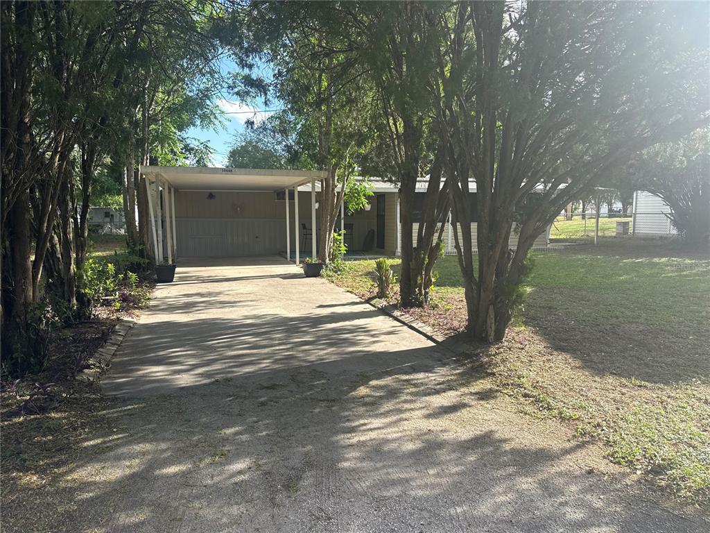 A single-story house features a concrete driveway leading to a covered carport under mature trees — 10448 SE 148TH PLACE, SUMMERFIELD, FL