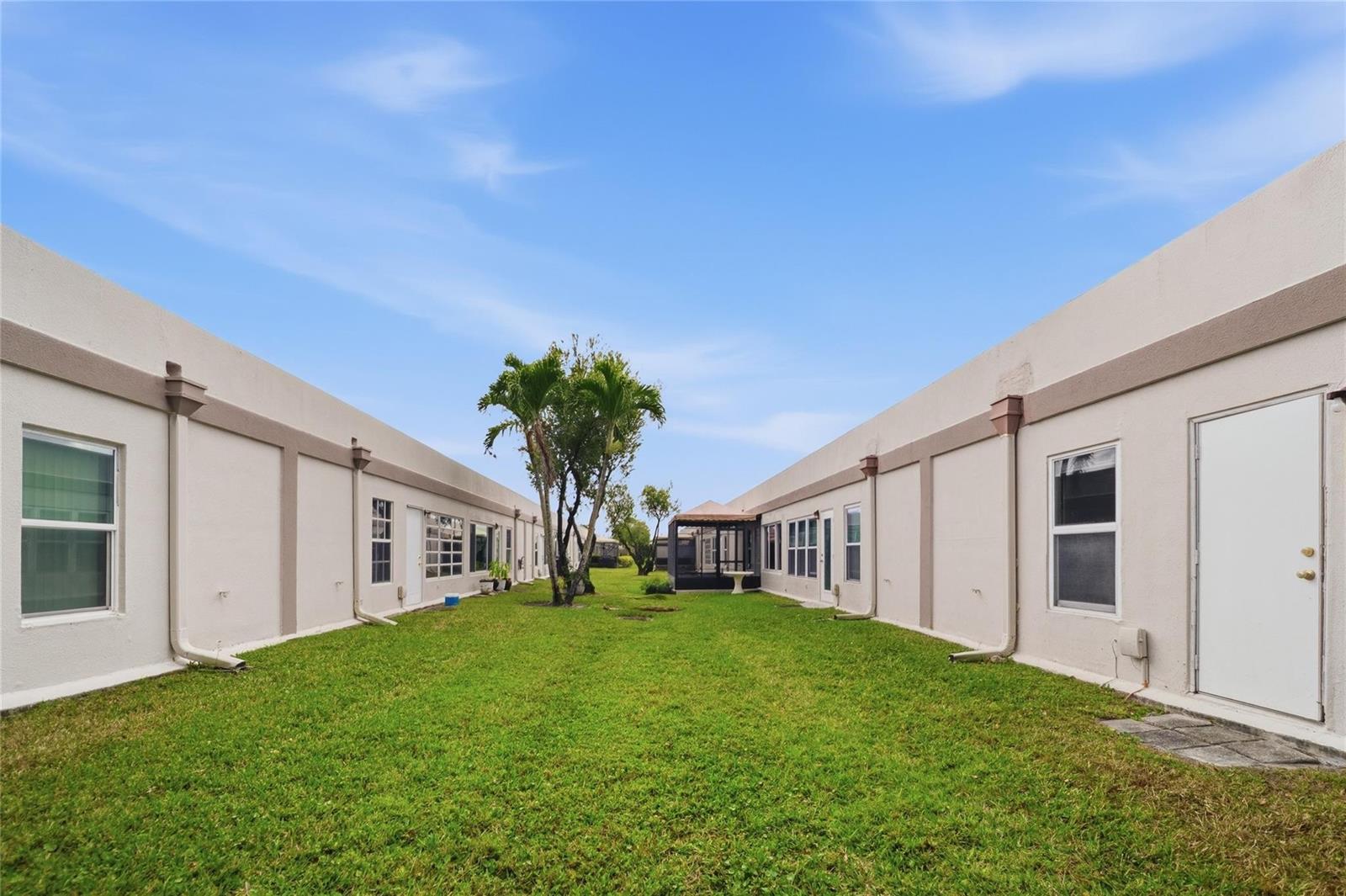 A grassy courtyard sits between two rows of single-story stucco buildings under a blue sky — 18730 Stewart Circle # 7, Boca Raton FL 33496, BOCA RATON, FL