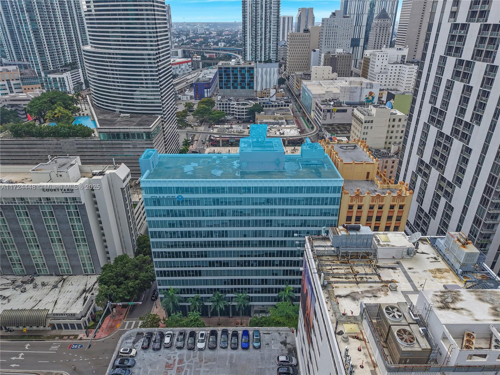 An aerial view shows a blue-tinted office building surrounded by other high-rise structures in a city — 150 SE 2nd Ave # 103, Miami FL 33131, MIAMI, FL