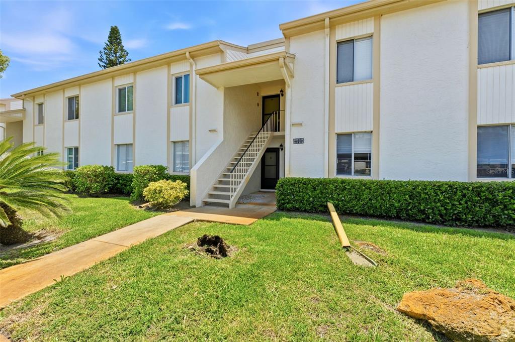 A two-story apartment building exterior features white stucco walls, a central staircase, and landscaping — 126 CYPRESS COURT UNIT 26, OLDSMAR, FL