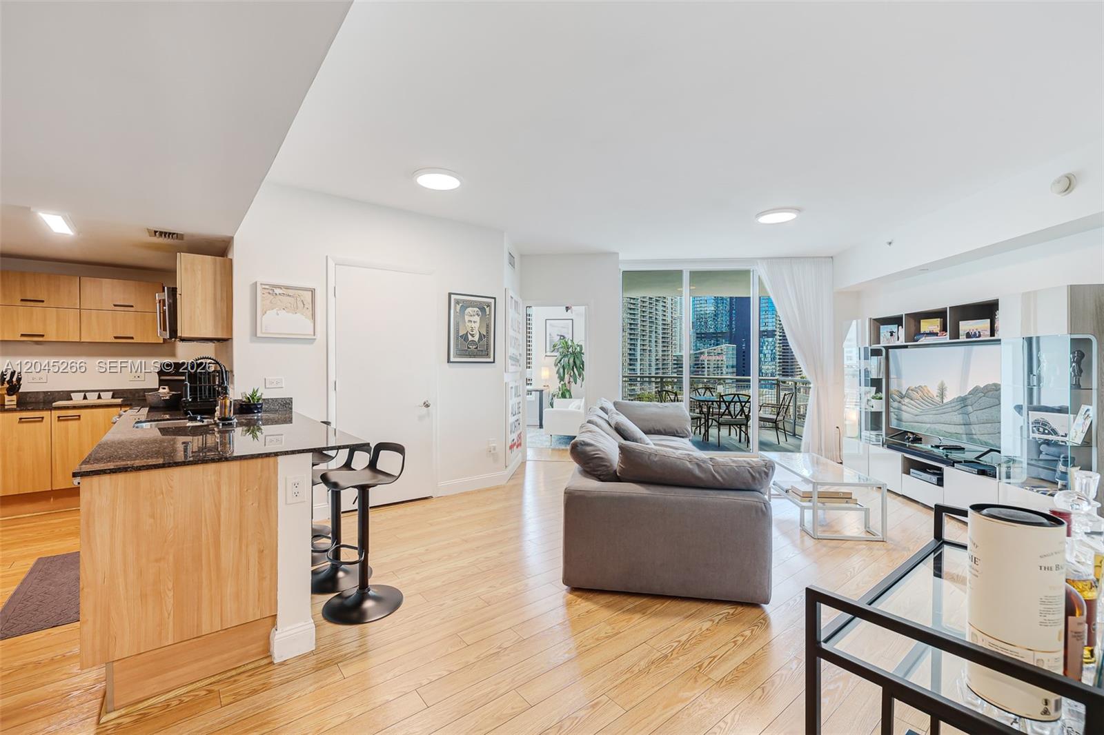A modern apartment features an open-concept kitchen with wood cabinets, a gray sofa, and balcony views — 350 S Miami Ave # 2104, Miami FL 33130, MIAMI, FL