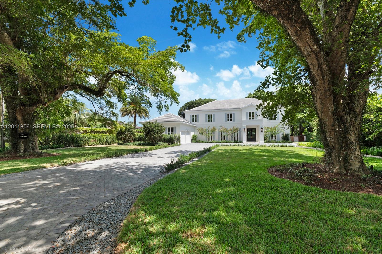 A two-story white house with a paved driveway sits on a grassy, tree-lined lot — 1241 Mariola Ct, Coral Gables FL 33134, CORAL GABLES, FL