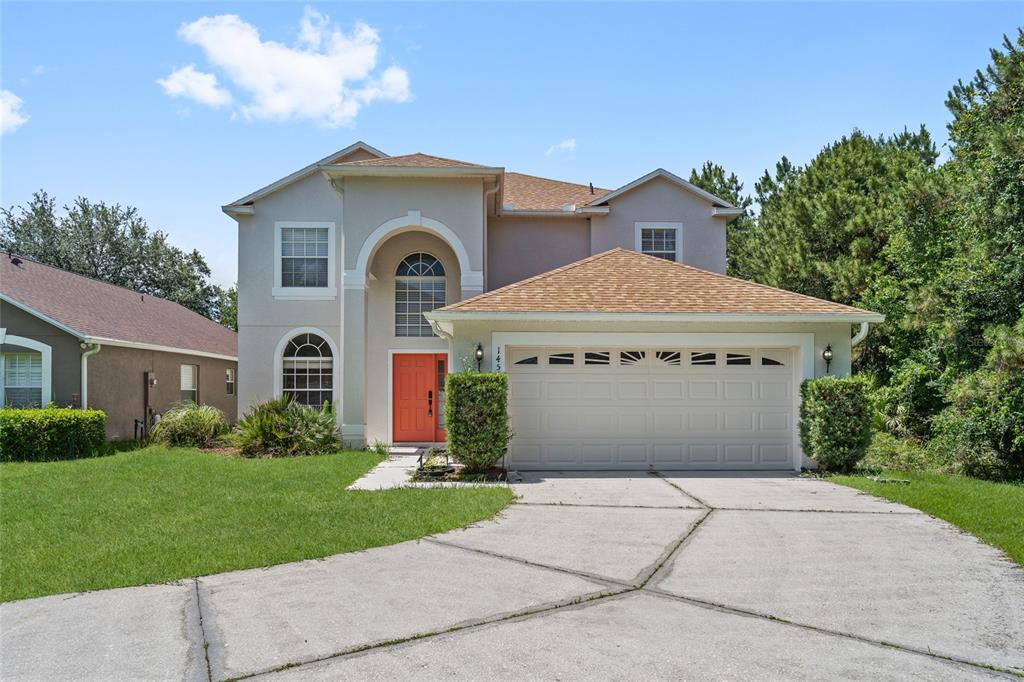 A two-story suburban house features a tan stucco exterior, a bright orange door, and driveway — 14539 UNBRIDLED DRIVE, ORLANDO, FL