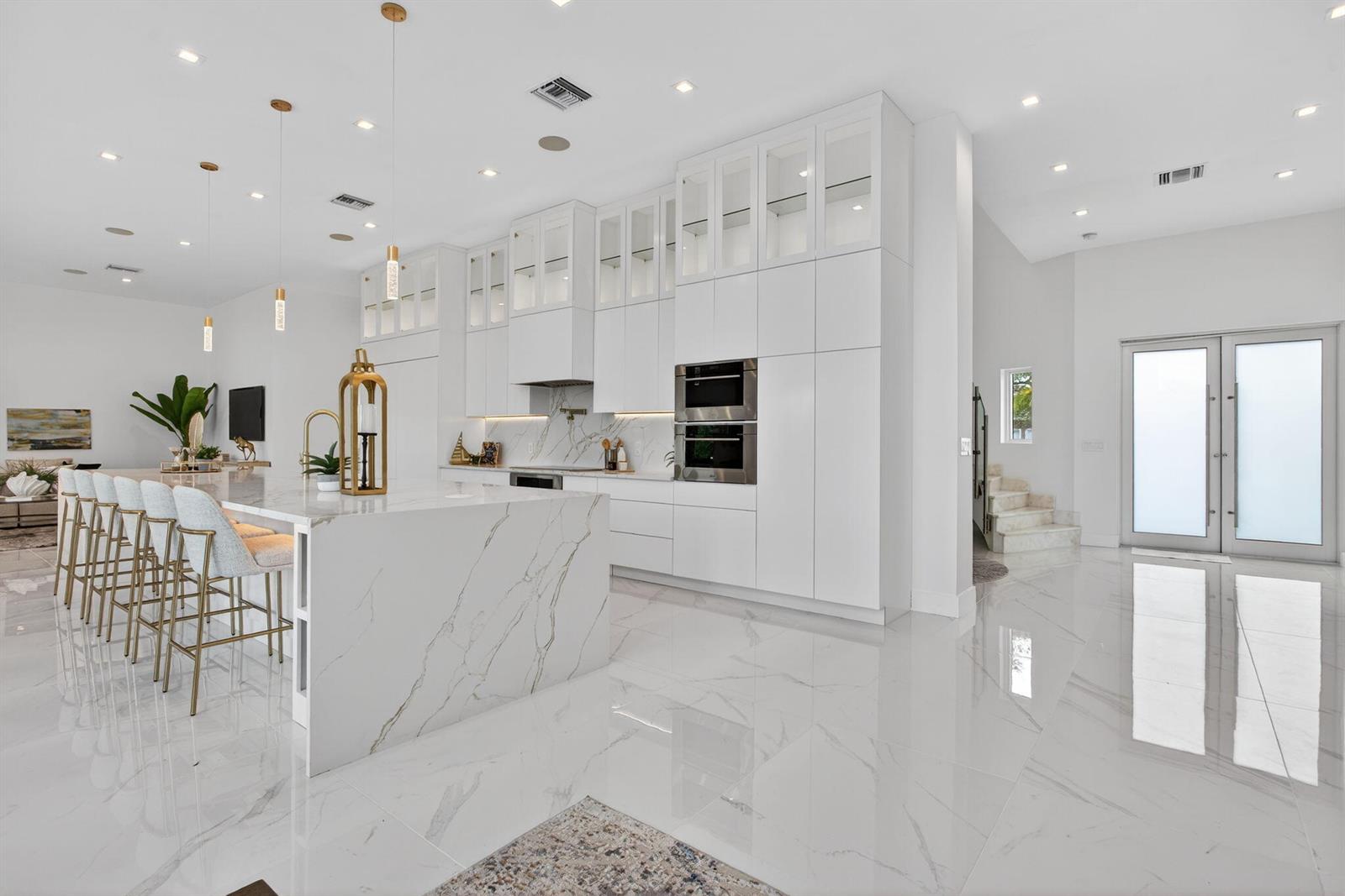A modern white kitchen features a marble waterfall island, gold bar stools, and glass-front cabinetry — 2641 NE 48th Street, Lighthouse Point FL 33064, LIGHTHOUSE POINT, FL