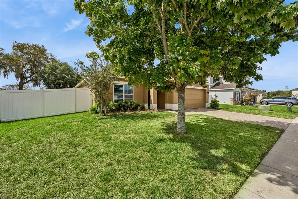 A tan single-story house with a two-car garage and a large tree in the yard — 240 WHISPERING PINES WAY, DAVENPORT, FL