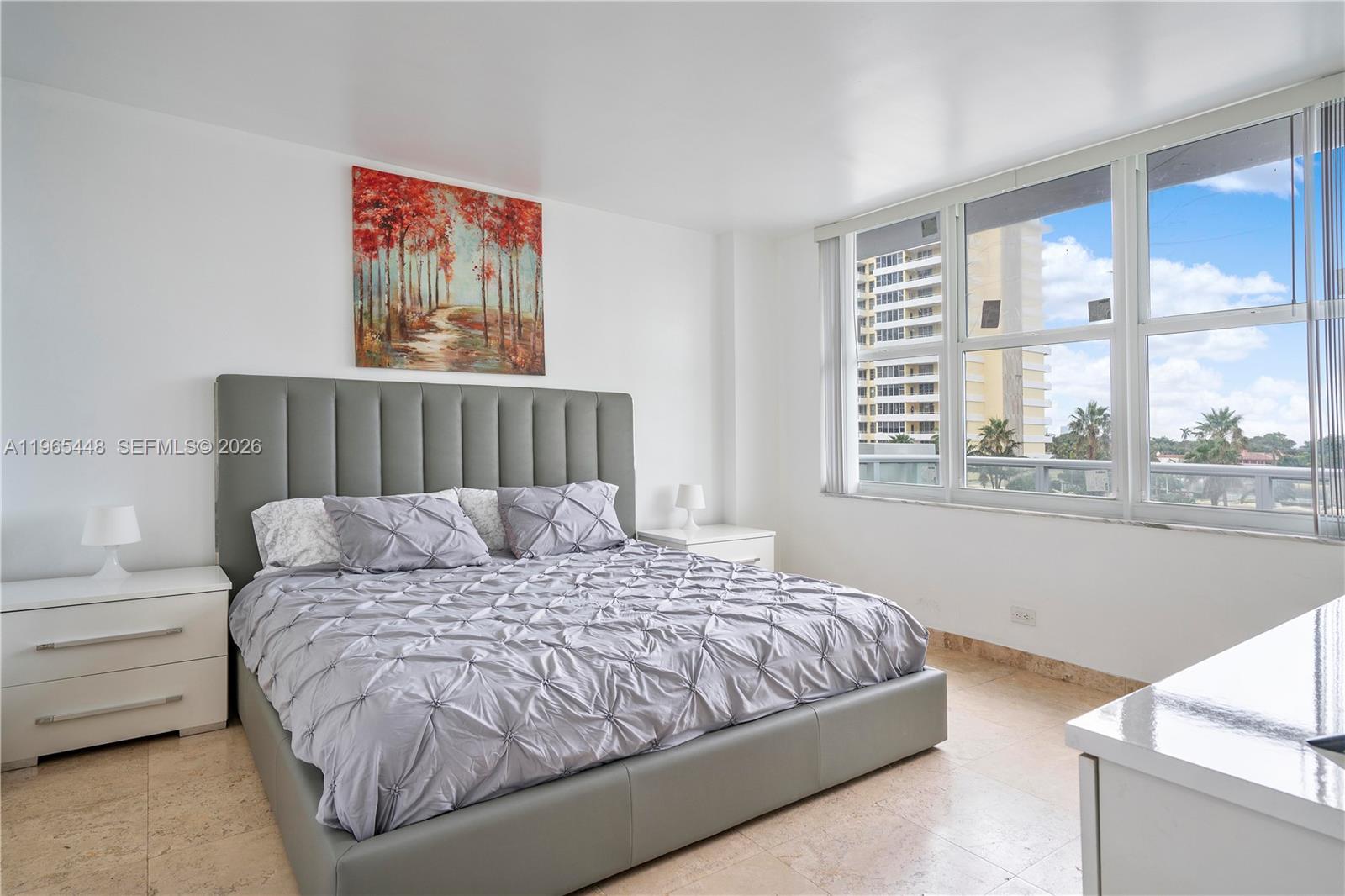 A bedroom features a gray upholstered bed, white nightstands, and large windows with city views — 5601 Collins Ave # 423, Miami Beach FL 33140, MIAMI BEACH, FL
