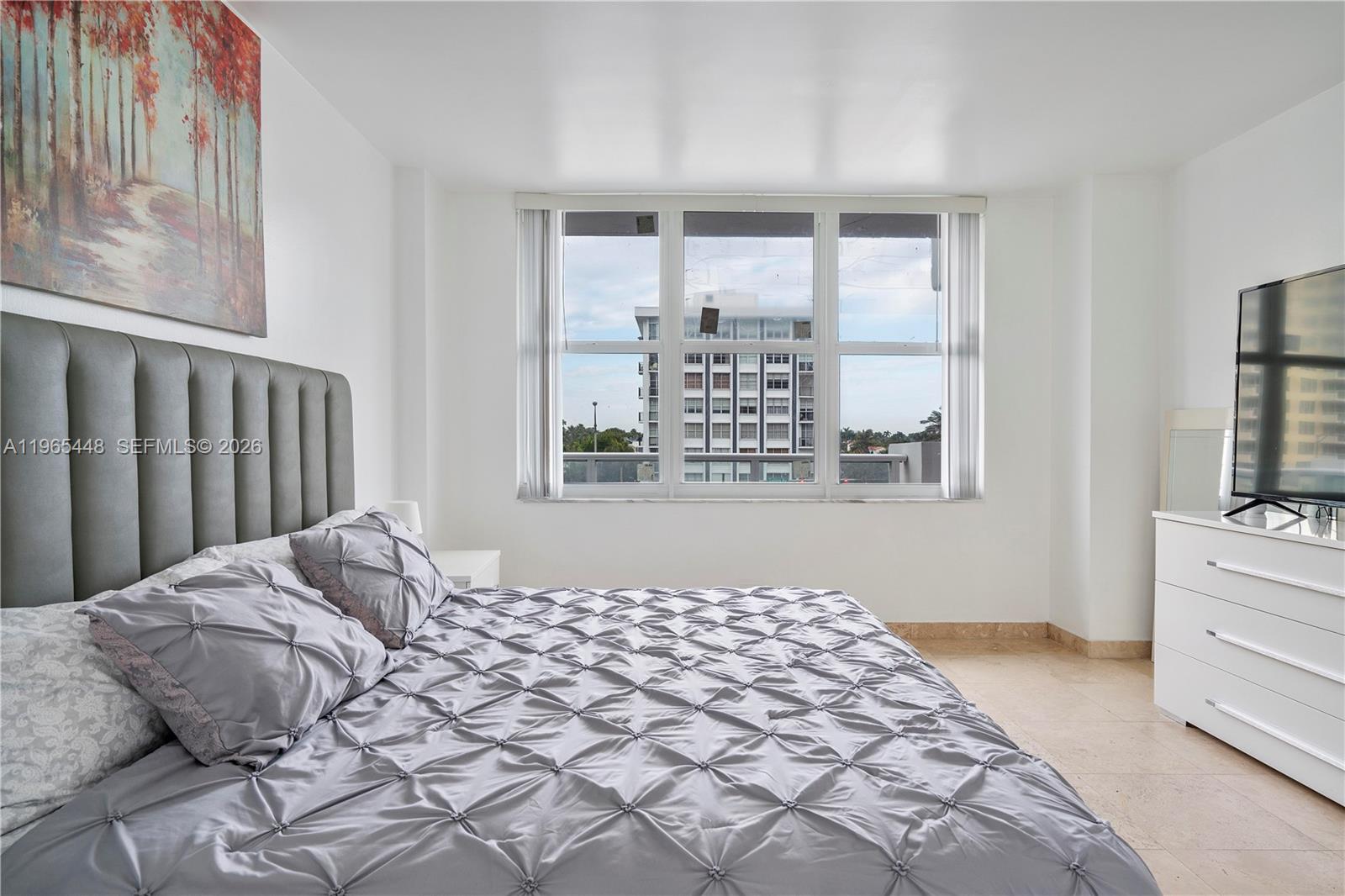 A bedroom features a grey tufted headboard, a white dresser, and a large window view — 5601 Collins Ave # 423, Miami Beach FL 33140, MIAMI BEACH, FL