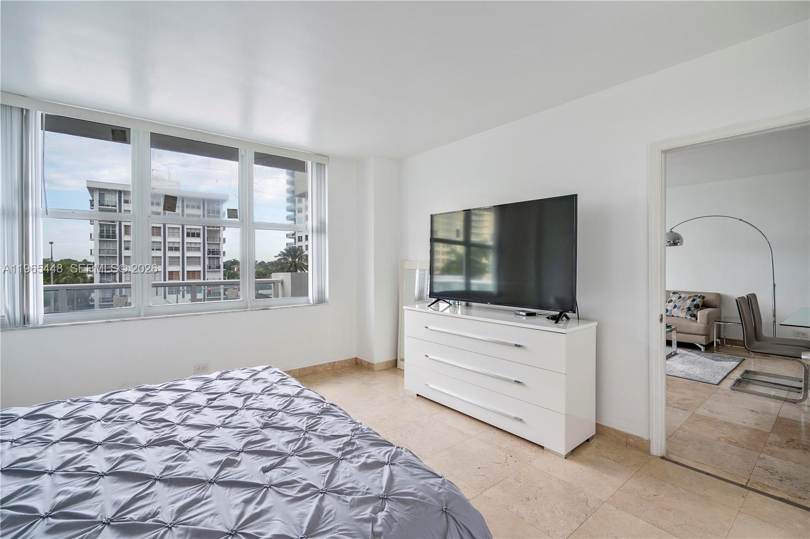 A bedroom features a gray tufted bed, a white dresser with a television, and tile flooring — 5601 Collins Ave # 423, Miami Beach FL 33140, MIAMI BEACH, FL