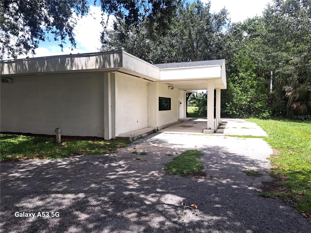 A white, flat-roofed commercial building features a covered drive-through area and a gravel parking lot — 6481 DELAND AVENUE, INDIAN LAKE ESTATES, FL