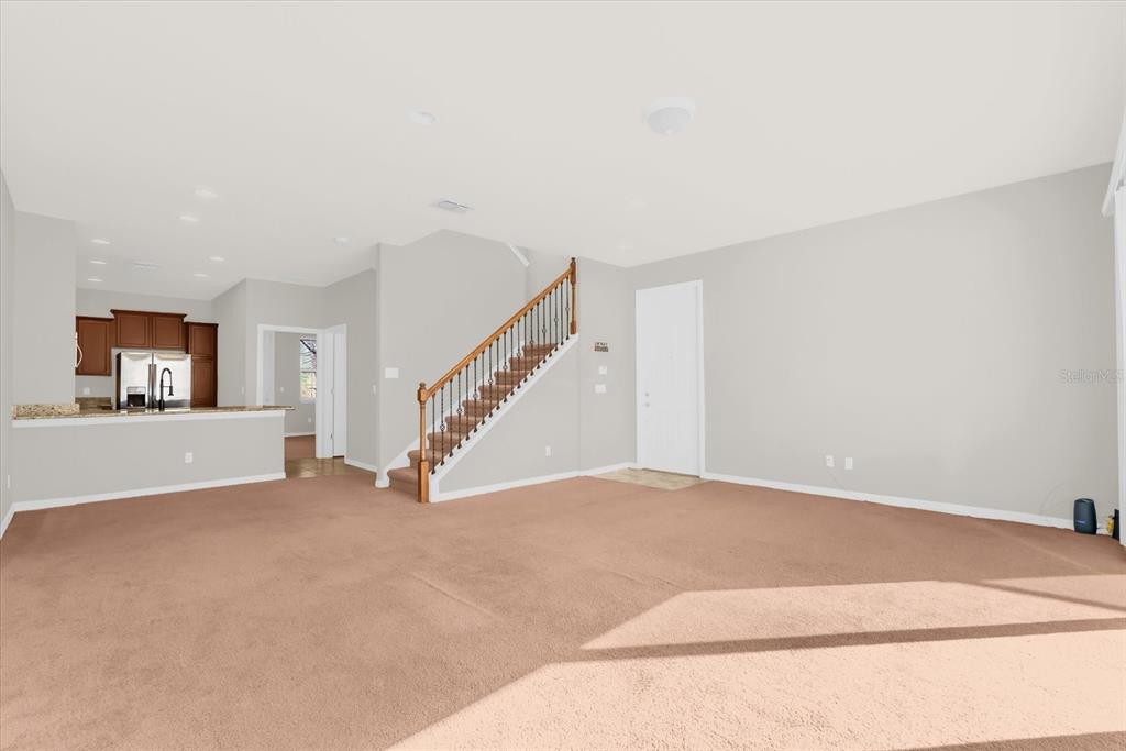An open-concept living room features tan carpeting, a wooden staircase, and an adjacent kitchen area — 11932 LORENZA LANE, ORLANDO, FL