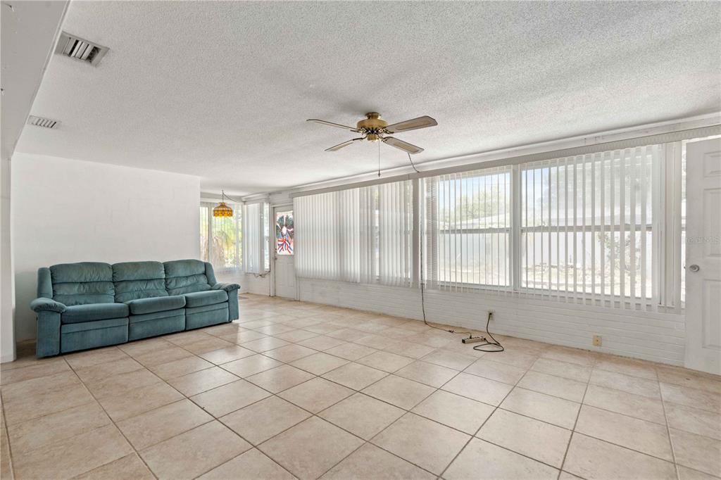 A sunroom features a teal sofa, tile flooring, a ceiling fan, and large vertical blinds — 927 PARKWOOD DRIVE, DUNEDIN, FL