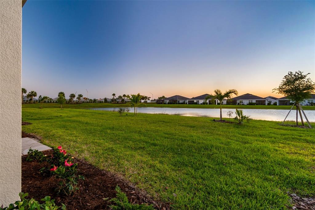 A view of a grassy backyard overlooking a pond with houses in the distance — 18122 BEACH RANCH TERRACE, LAKEWOOD RANCH, FL