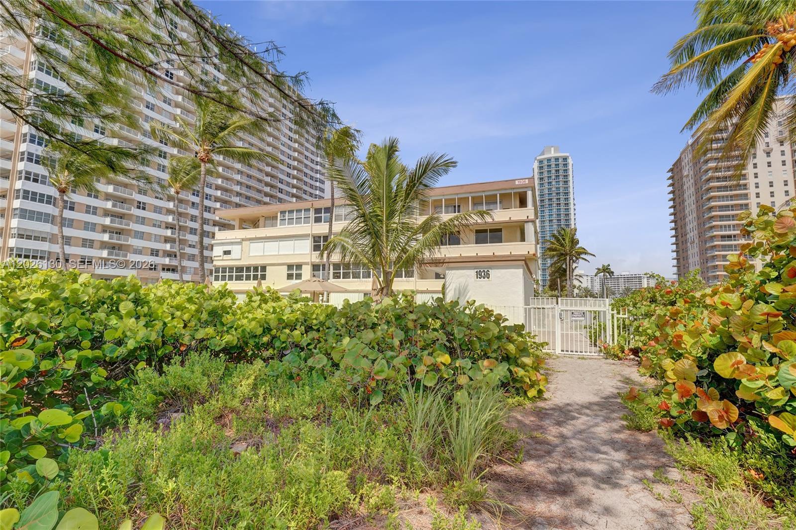 A low-rise building with a gated entrance is surrounded by lush greenery and tall towers — 1936 S Ocean Dr # 4C, Hallandale Beach FL 33009, HALLANDALE BEACH, FL