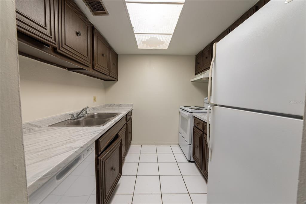 A galley kitchen features dark wood cabinets, white appliances, marble-look countertops, and white tile flooring — 4718 JOSEPH COURT UNIT 220, TAMPA, FL