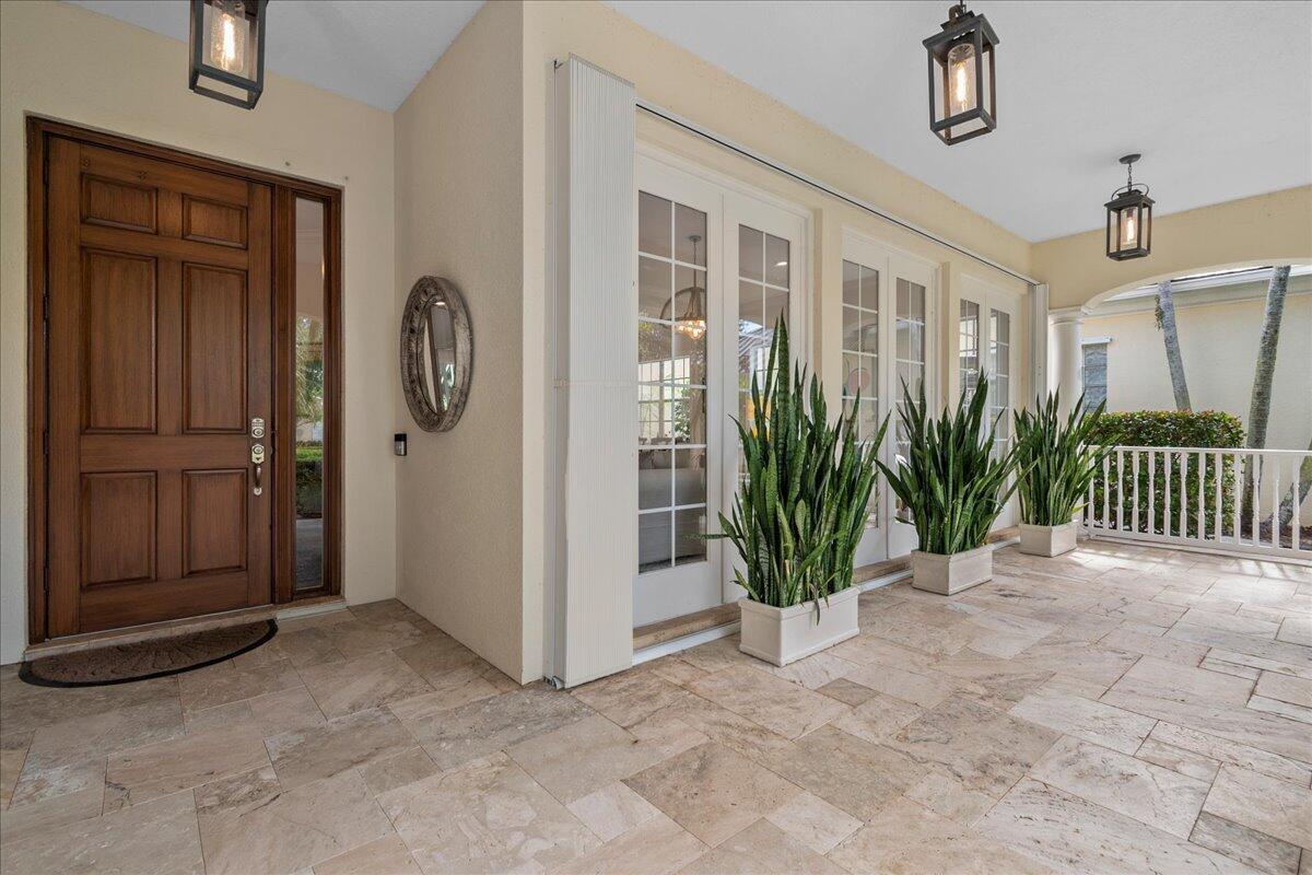 A covered patio features travertine tile flooring, a wooden front door, and potted snake plants — 8465 Belize Place, Wellington FL 33414, WELLINGTON, FL