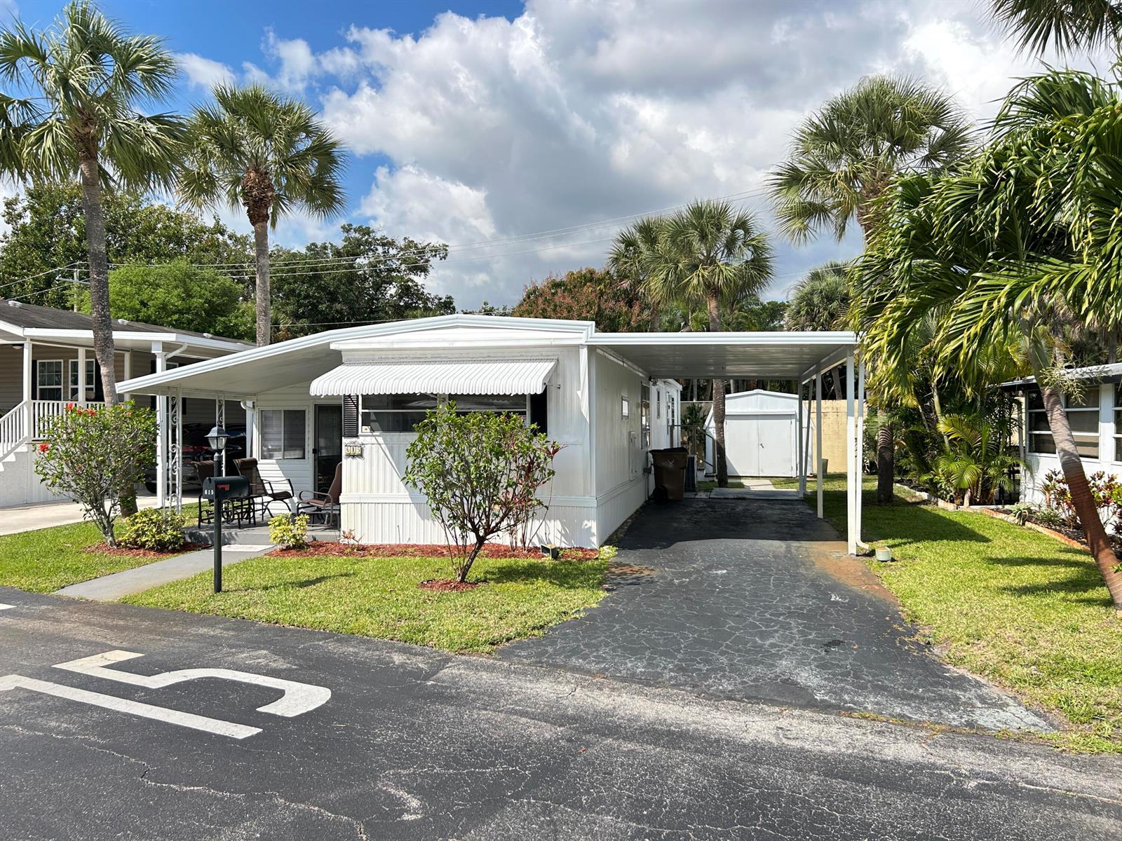A white mobile home with a carport, front porch, and palm trees under a cloudy sky — 615 Alamanda Lane, Deerfield Beach FL 33442, DEERFIELD BEACH, FL