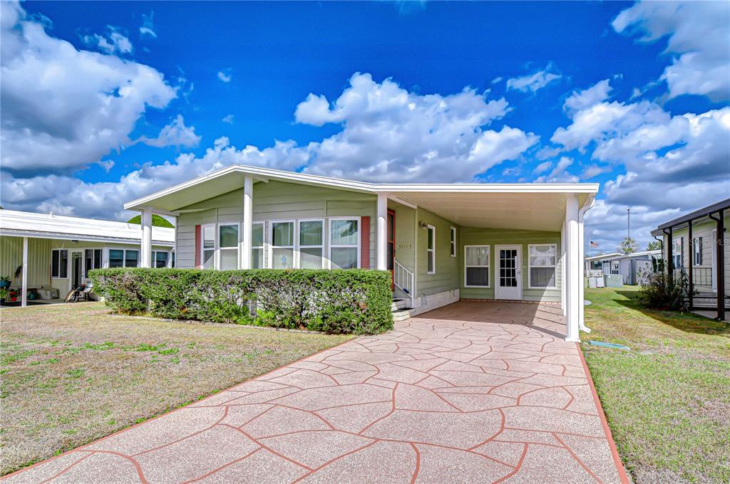 A light green manufactured home features a paved driveway, a carport, and a front hedge — 35145 WAGNER WAY, ZEPHYRHILLS, FL