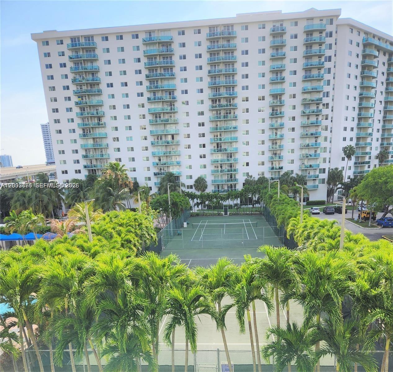 An exterior view shows a tall apartment building overlooking two tennis courts and palm trees — SUNNY ISLES BEACH, FL