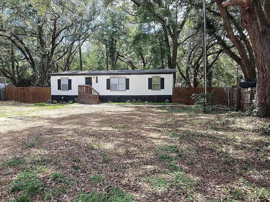 A single-story manufactured home sits on a dirt lot surrounded by mature trees and fencing — 6626 NW 60 CT #9, OCALA, FL