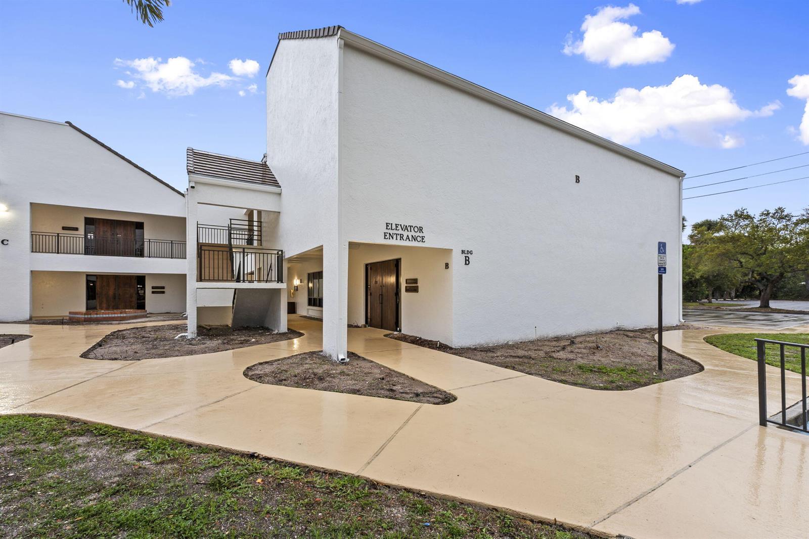 The exterior of a white multi-story building features an elevator entrance and a paved walkway — 900 SE Ocean Boulevard # B212, Stuart FL 34994, STUART, FL