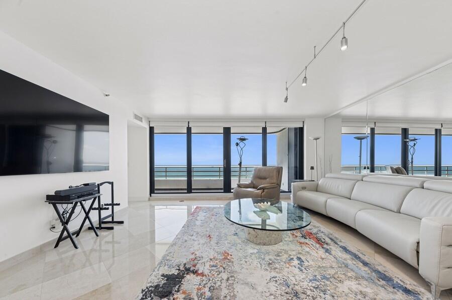 A living room with a large television, white sofa, glass coffee table, and ocean view — 1400 S Ocean Boulevard # 1004, Boca Raton FL 33432, BOCA RATON, FL