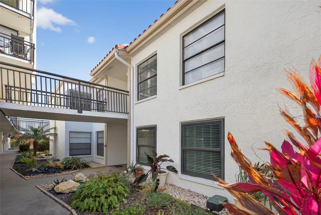 A two-story building exterior features a pedestrian bridge, stucco walls, and a landscaped garden — 6210 SUN BOULEVARD UNIT 112, ST PETERSBURG, FL