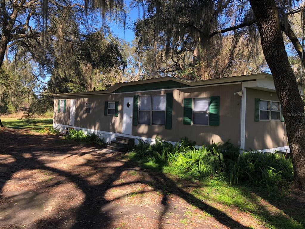 A tan mobile home with green shutters sits under large trees with hanging Spanish moss — 6382 BAKER ROAD, KEYSTONE HEIGHTS, FL