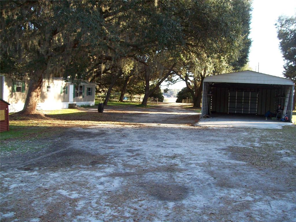 A mobile home and a detached metal carport sit on a large, tree-shaded dirt lot — 6382 BAKER ROAD, KEYSTONE HEIGHTS, FL