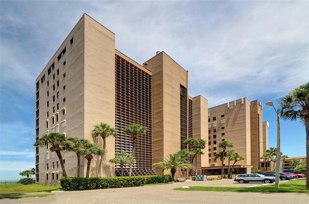 A tall, tan, multi-story apartment building stands behind a parking lot with palm trees — 900 GULF BOULEVARD UNIT 907, INDIAN ROCKS BEACH, FL