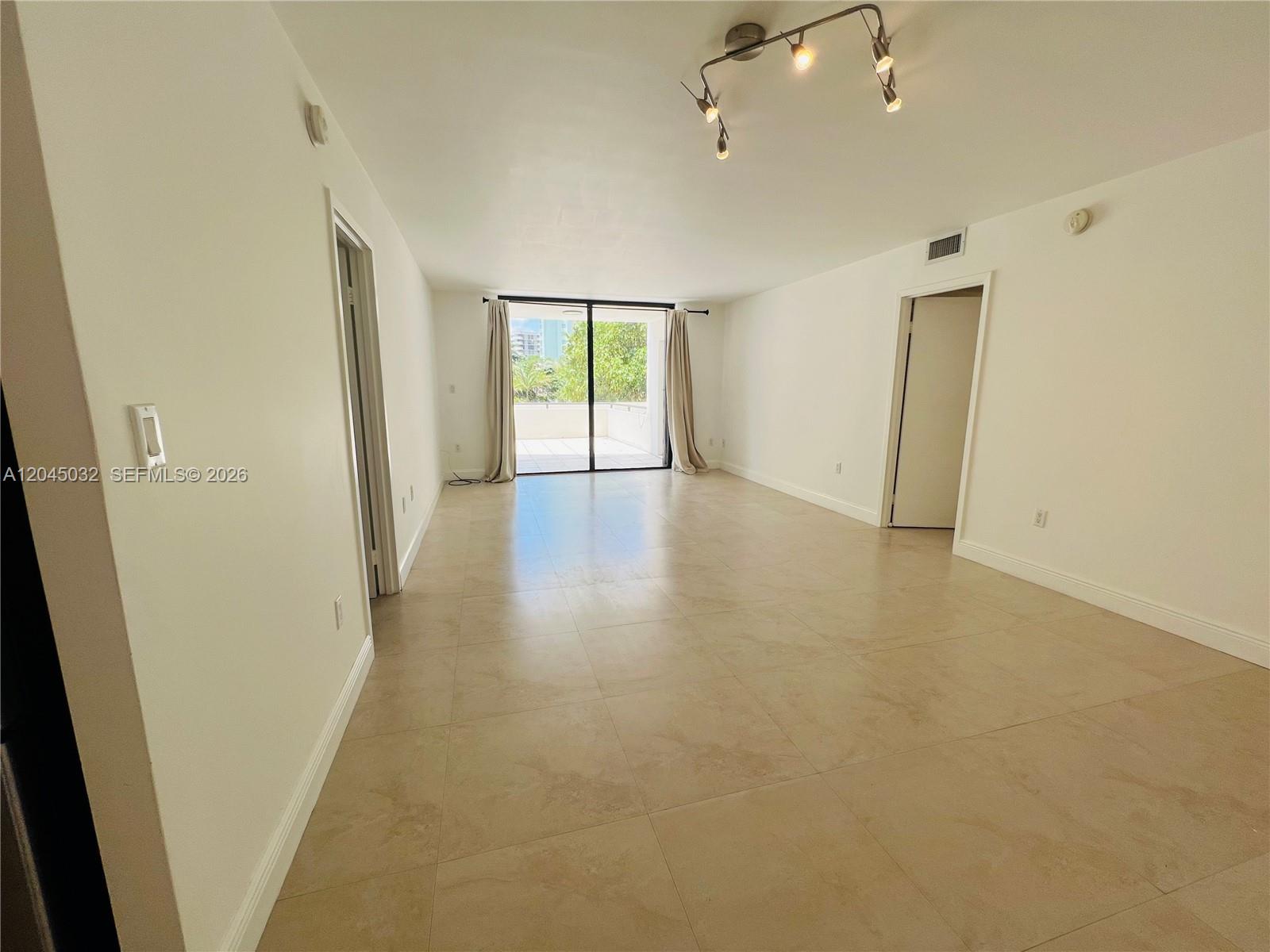 An empty living room features beige tile flooring, white walls, and a sliding glass balcony door — 2333 Brickell Ave # 210, Miami FL 33129, MIAMI, FL